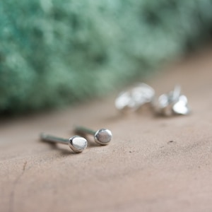 Può includere: Un paio di piccoli orecchini a perno in argento con una forma arrotondata leggermente irregolare. Gli orecchini sono su una superficie marrone.