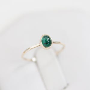 Può includere: Un delicato anello d'oro con una piccola gemma verde smeraldo di forma ovale. L'anello ha una sottile fascia d'oro. La gemma è incastonata in una semplice lunetta, che ne evidenzia il colore vibrante.