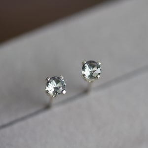 May include: A pair of stud earrings featuring round, faceted, pale blue gemstones set in silver-colored metal. The earrings are displayed on a light gray surface, showcasing their delicate design and sparkle.