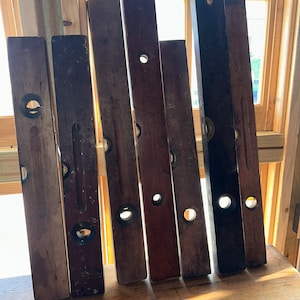May include: A collection of vintage wooden levels, each with a dark, aged appearance. The levels are arranged vertically, showcasing their rectangular shape and circular bubble vials. The wood tones vary from light to dark brown, suggesting age and use.