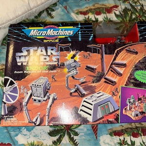 May include: A Micro Machines Space Star Wars Endor playset from Return of the Jedi. The box features a red and white AT-ST walker, a gray bunker, and a brown log ramp. The playset includes Super Space Action Features.