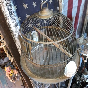May include: A vintage brass birdcage with a wooden perch and a white ceramic bird figurine inside. The cage is hanging from a chain and has a round base.