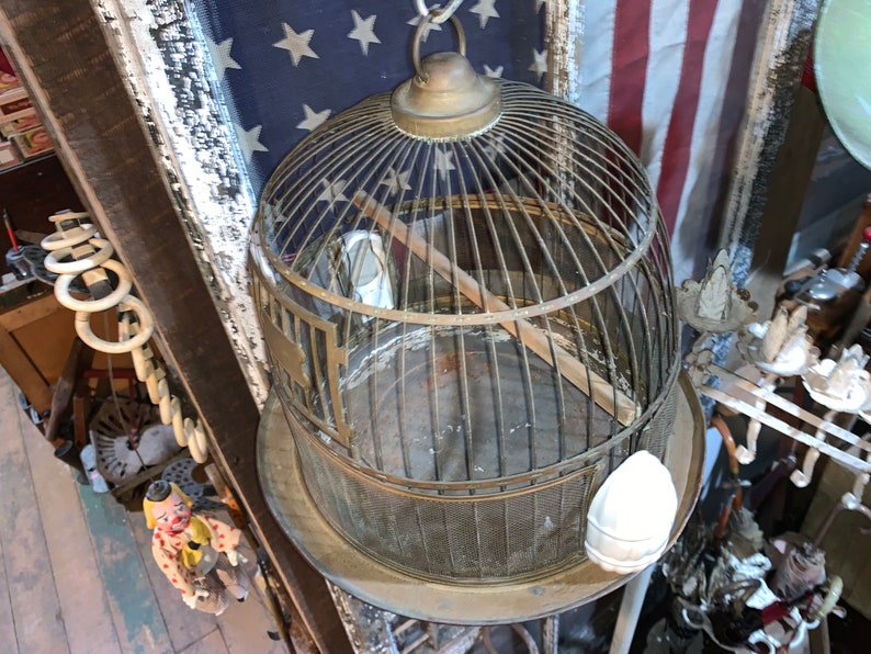 May include: A vintage brass birdcage with a wooden perch and a white ceramic bird figurine inside. The cage is hanging from a chain and has a round base.