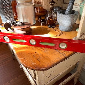 May include: A red wooden level with three green bubble vials. The level is lying on a wooden table.