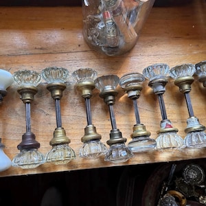 May include: A collection of nine door knobs arranged in a row. One knob is opaque white glass, while the other eight are clear, faceted glass. Each knob features a metal spindle and brass or metallic hardware components connecting the two ends.