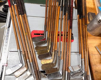 May include: A collection of vintage golf clubs with wooden shafts and various club heads. The grips are wrapped in leather or other materials, and the clubs show signs of age and use. The clubs are arranged on a light-colored surface.