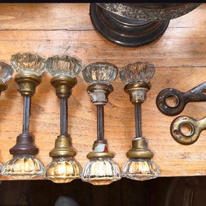 May include: Five clear glass door knobs with fluted designs and brass-toned metal bases, each featuring a long metal spindle. Two separate teardrop-shaped metal escutcheon plates with keyholes are positioned to the right of the door knobs.