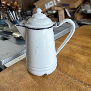 May include: White enamel coffee pot with a lid and handle. The pot has a blue trim around the spout and lid. The pot is sitting on a wooden surface.