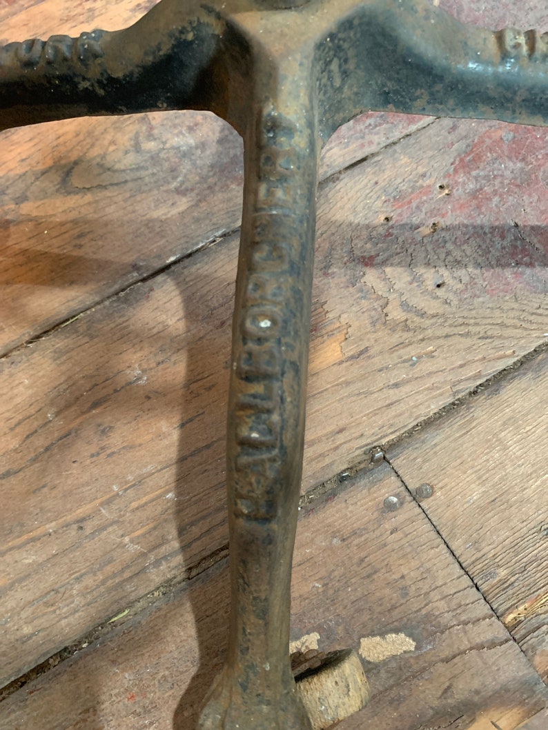 以下が含まれることがあります： 「The Goshen Mfg. Co.」という文字が側面に刻印された鋳鉄製の土台をクローズアップ。土台は3本の脚で構成され、表面は粗くてざらざらしている。