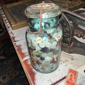 May include: A clear glass jar filled with a variety of buttons in different colors and sizes. The jar has a glass lid and a metal handle. The buttons are a mix of white, cream, and other colors. The jar is sitting on a white distressed table.