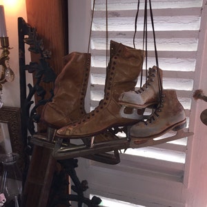 May include: A pair of brown leather ice skates with metal blades hanging from a white slatted window. The skates have laces and are in good condition.