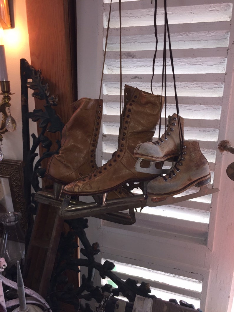 May include: A pair of brown leather ice skates with metal blades hanging from a white slatted window. The skates have laces and are in good condition.