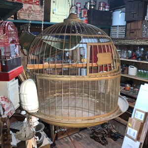 May include: A large, antique, brass birdcage with a round, domed top and a wire mesh bottom. The cage is hanging from a chain and is surrounded by other antique items.