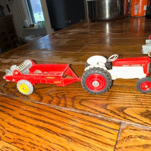 May include: A red and white toy tractor with a red wagon attached. The tractor has the words "Massey Ferguson 65" printed on the side. The wagon has the words "Massey Harris" printed on the side.