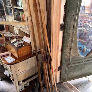 May include: A collection of vintage wooden bows leaning against a weathered wooden door frame. The bows are various shades of brown with dark red and tan accents. The scene is set in a rustic, antique-filled room with a distressed white cabinet and a glass-paned door.