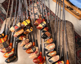 May include: A collection of vintage golf clubs with wooden heads and steel shafts. The club heads feature various wood grains, painted accents in red, black, and yellow, and decorative inlay patterns. The shafts are thin, metallic, and silver-toned.