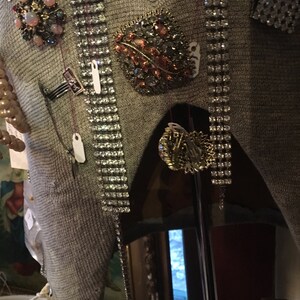 May include: A gray sweater with a variety of brooches and pins, including a large, ornate brooch with a leaf design and rhinestones. The sweater is displayed on a mannequin.