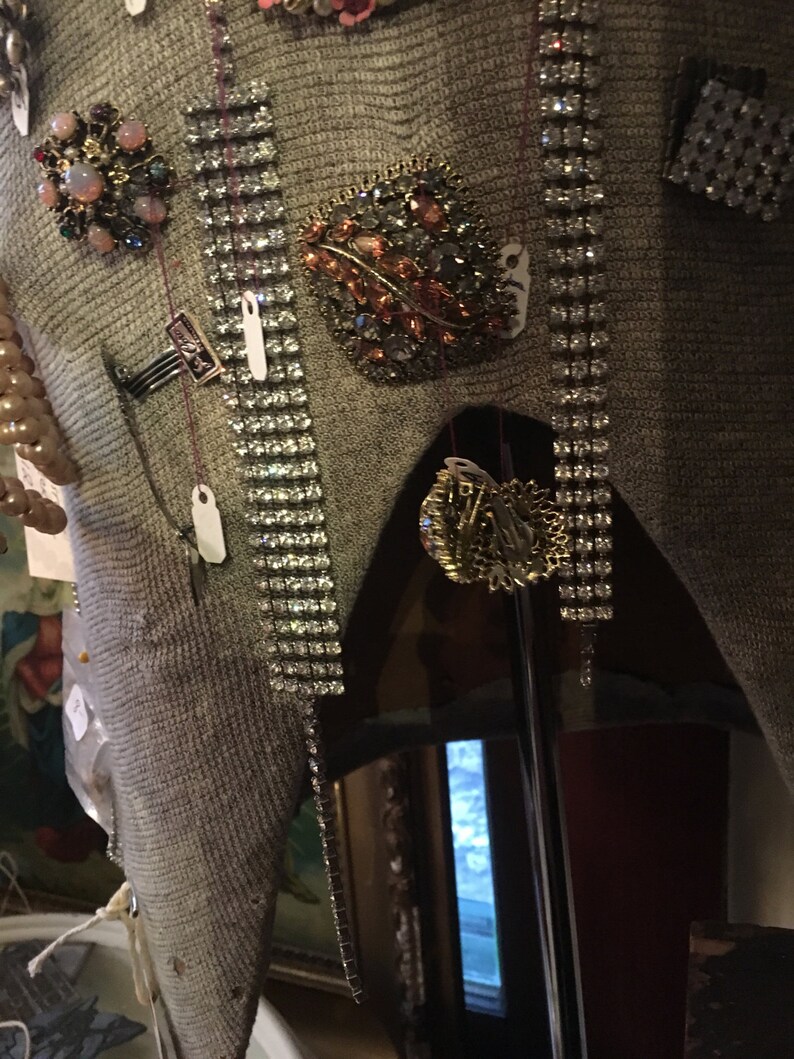 May include: A gray sweater with a variety of brooches and pins, including a large, ornate brooch with a leaf design and rhinestones. The sweater is displayed on a mannequin.