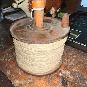 May include: A vintage wooden spool with a brown metal base and a wooden top. The spool is filled with beige twine and has a small brown plastic cap on the side.