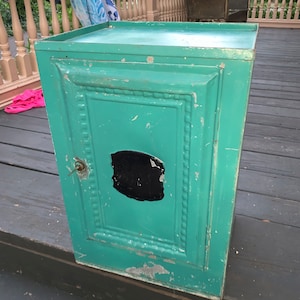 May include: A teal green metal cabinet with a single door and a black label. The cabinet has a decorative design around the door and a small keyhole.