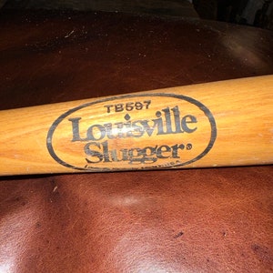 May include: A wooden Louisville Slugger baseball bat with a black oval label that reads "Louisville Slugger" and "TB597".