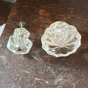 May include: Two clear, faceted glass perfume bottle components sit on a dark wooden surface. One is a stopper with a metal stem, the other is a round, domed bottle top with a starburst design.