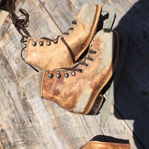 May include: A pair of vintage leather ice skates with brown laces and metal eyelets. The skates have a worn, distressed look and are sitting on a wooden surface.