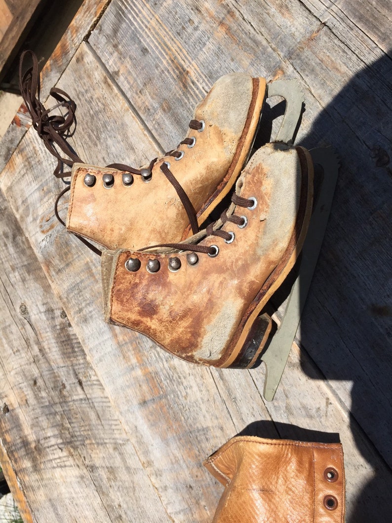 May include: A pair of vintage leather ice skates with brown laces and metal eyelets. The skates have a worn, distressed look and are sitting on a wooden surface.