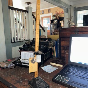 May include: A wooden ruler with a black base and a small glass jar hanging from it. The ruler is marked with numbers from 1 to 20. The ruler is sitting on a wooden table with a laptop, a typewriter, and other items.