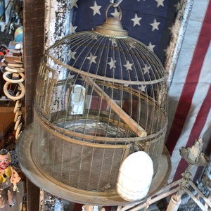 May include: A vintage brass birdcage with a wire mesh design hangs from a chain. The cage has a round top and a flat bottom. The cage is empty and has a wooden perch inside. The cage is hanging in front of an American flag.