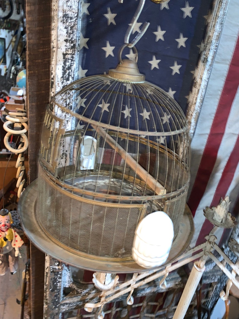May include: A vintage brass birdcage with a wire mesh design hangs from a chain. The cage has a round top and a flat bottom. The cage is empty and has a wooden perch inside. The cage is hanging in front of an American flag.
