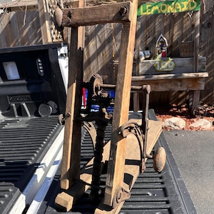 May include: An antique wooden apple press with metal gears and a screw mechanism. The press is made of weathered wood and has a rustic appearance. The press is sitting on the bed of a truck.