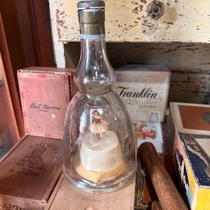 May include: A clear glass bottle with a ballerina figurine inside, resting on a white base. The bottle has a metal cap and is surrounded by vintage cigar boxes, including one labeled "Franklin Cigars."