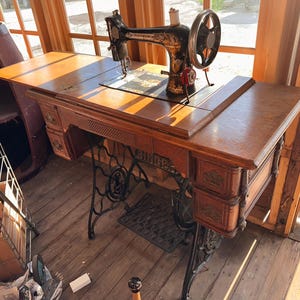 May include: A vintage Singer sewing machine mounted on a wooden cabinet with a fold-out top. The machine is black with gold decorative decals and a large hand wheel. The cabinet features multiple drawers with ornate metal pulls and a treadle base.