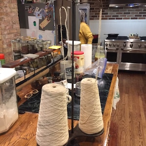 May include: Two spools of beige yarn are wound on a metal yarn winder. The winder is on a wooden table with a green marble surface. The yarn is ready to be used for knitting or crocheting.