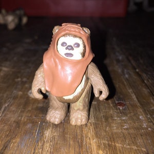 May include: A brown Ewok action figure from the Star Wars franchise. The figure has a tan face, brown fur, and wears a brown hood. The figure is standing on a wooden surface.