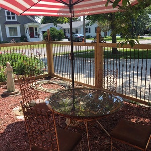 May include: A red and white striped patio umbrella provides shade over a glass-top table and four rusty metal chairs. The table and chairs are set on a bed of brown wood chips.