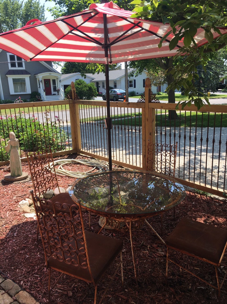 May include: A red and white striped patio umbrella provides shade over a glass-top table and four rusty metal chairs. The table and chairs are set on a bed of brown wood chips.
