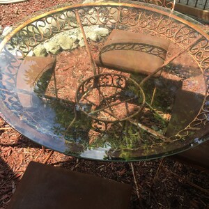 May include: A rusty metal patio table with a glass top and a decorative scrolled design. The table is surrounded by wood chips.