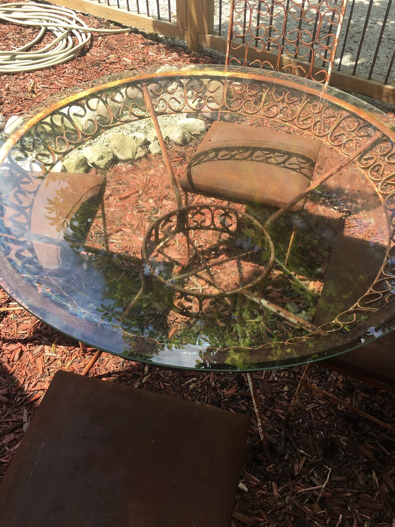 May include: A rusty metal patio table with a glass top and a decorative scrolled design. The table is surrounded by wood chips.