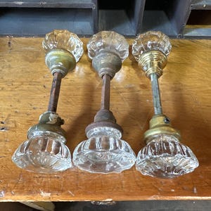 May include: Three vintage door knobs featuring clear, faceted glass handles with a fluted, starburst-like pattern. Each knob is attached to a central metal spindle and includes a circular brass-toned base plate with visible screws and aged patina.