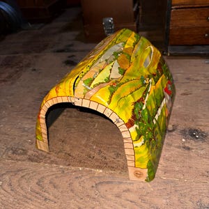 May include: A colorful toy train tunnel with a painted landscape scene. The tunnel is arched with a tan brick-like design at the entrance. The landscape features green hills, trees, and a yellow sky.