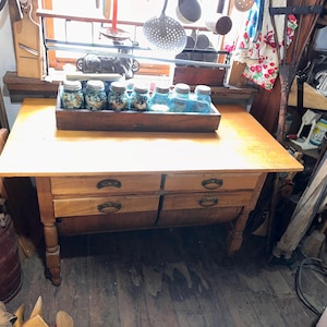 May include: A light brown wooden table featuring four drawers with dark metal pulls. A rectangular wooden tray sits on the tabletop, containing six blue glass jars filled with various small items. The table has four sturdy legs.