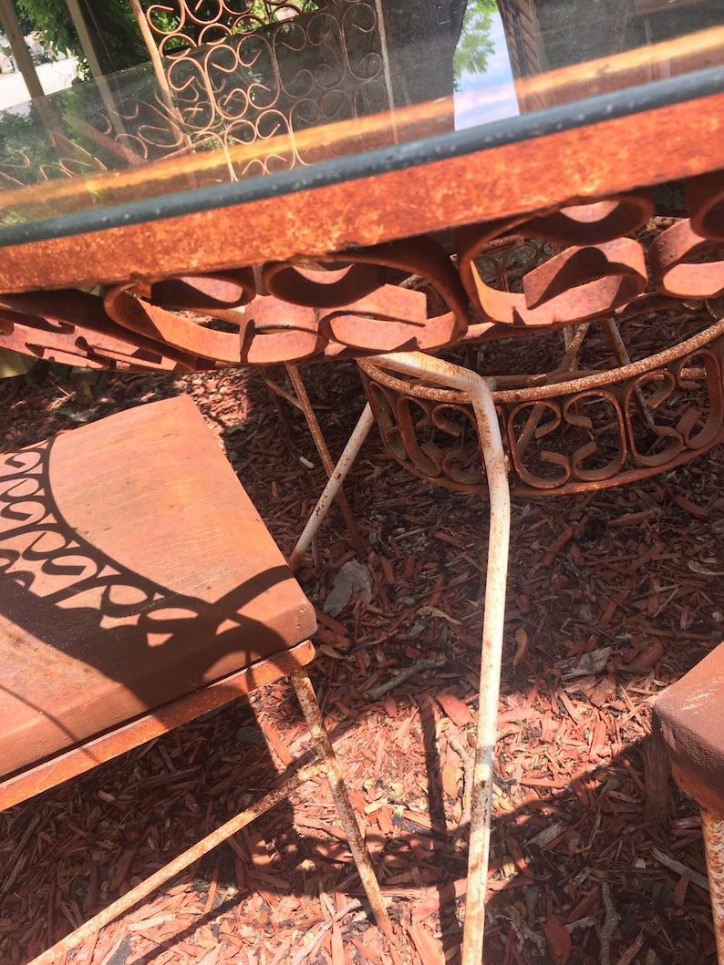 May include: A rusty metal patio table set with a glass top and white painted legs. The table and chairs are sitting on a bed of red wood chips.