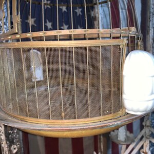 May include: A vintage brass birdcage with a wire mesh bottom and a white ceramic birdhouse attached to the side.