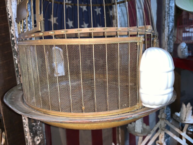May include: A vintage brass birdcage with a wire mesh bottom and a white ceramic birdhouse attached to the side.