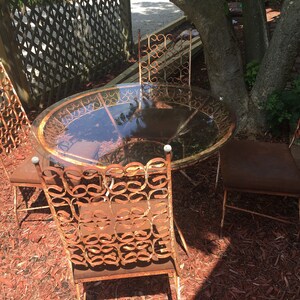 May include: A rusty metal patio set with a round glass table and four chairs. The chairs have intricate, ornate designs and are surrounded by red wood chips.