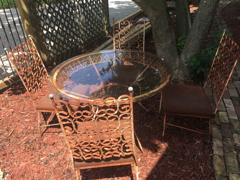 May include: A rusty metal patio set with a round glass table and four chairs. The chairs have intricate, ornate designs and are surrounded by red wood chips.