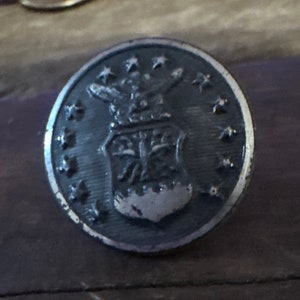 May include: A silver-colored button with a design of a shield and eagle with stars around the edge.