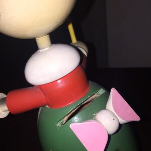 以下が含まれることがあります： 大きな頭と緑色の体を持つフィギュアの形をした、ヴィンテージの木製おもちゃの銀行。フィギュアは赤いスカーフ、白い帽子、ピンクのリボンをしています。銀行にはコインを挿入するためのスロットがあります。